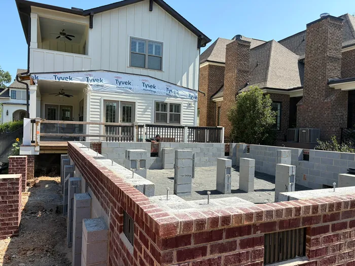 Brick-faced block foundation walls and concrete block piers with anchor bolts for the addition, beside the white home with Tyvek wrap along its first-floor wall