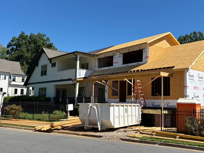 Addition framing going up beside the existing white farmhouse-style home: sheathed walls and roof decking, ladders, lumber stacked at the curb and a roll-off dumpster