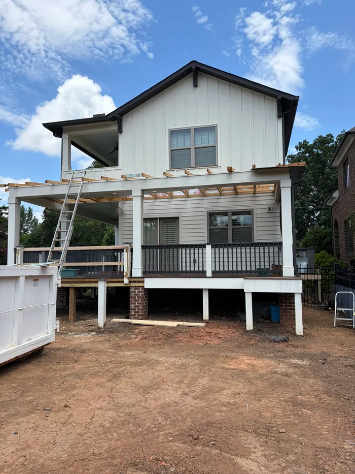Rear of the white farmhouse-style home with its pergola roof opened to bare rafters, a ladder leaning on the porch, a roll-off dumpster and a bare dirt yard