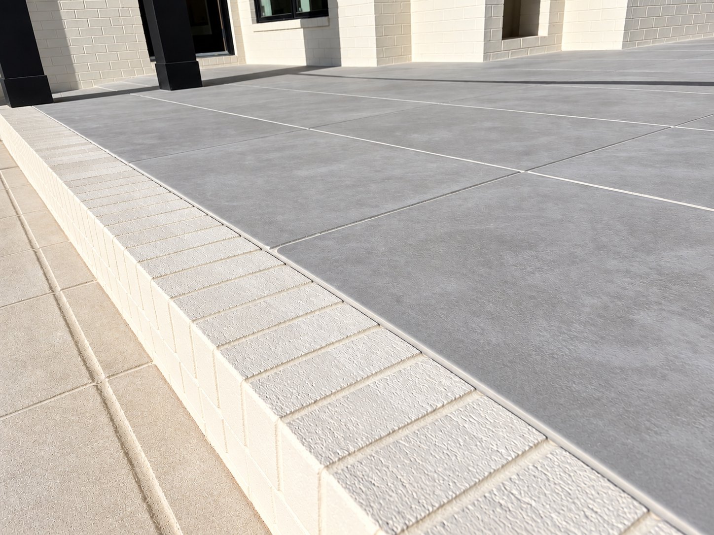 A finished elevated patio where large gray pavers meet a cream brick rowlock edge, beside light brick walls and black porch columns.