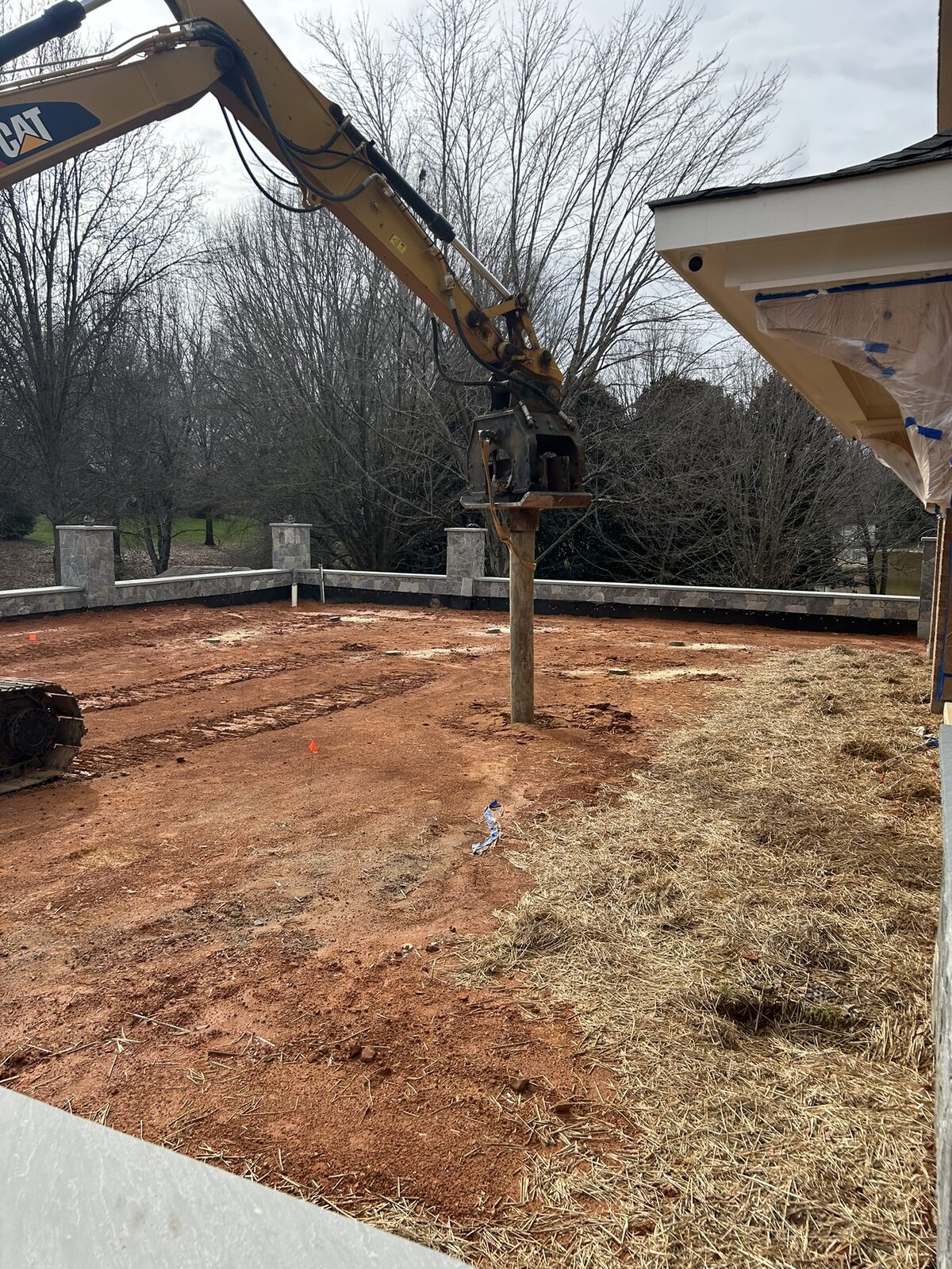 An excavator driving a piling into the motor court pad ahead of the structural concrete waffle slab, with the finished stone retaining wall behind.