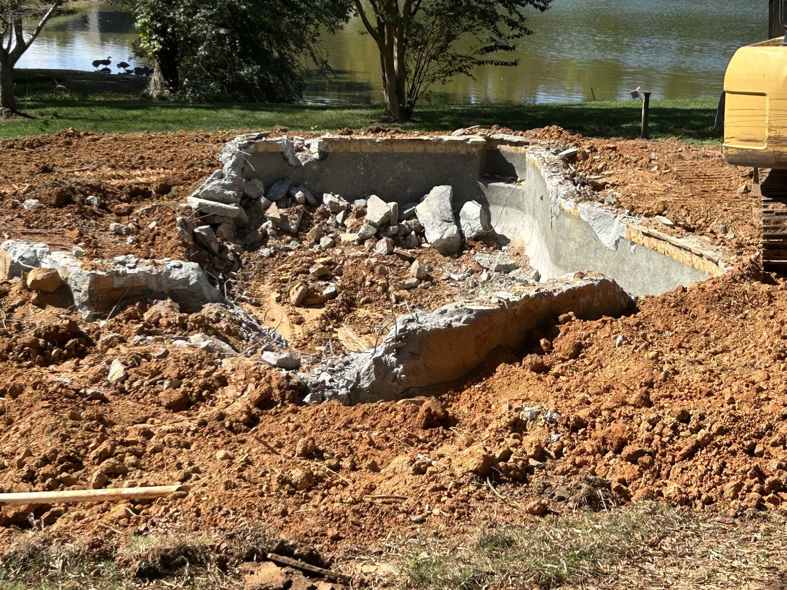 The original in-ground pool broken apart by an excavator, concrete shell and rubble sitting in red clay with the lake behind it.