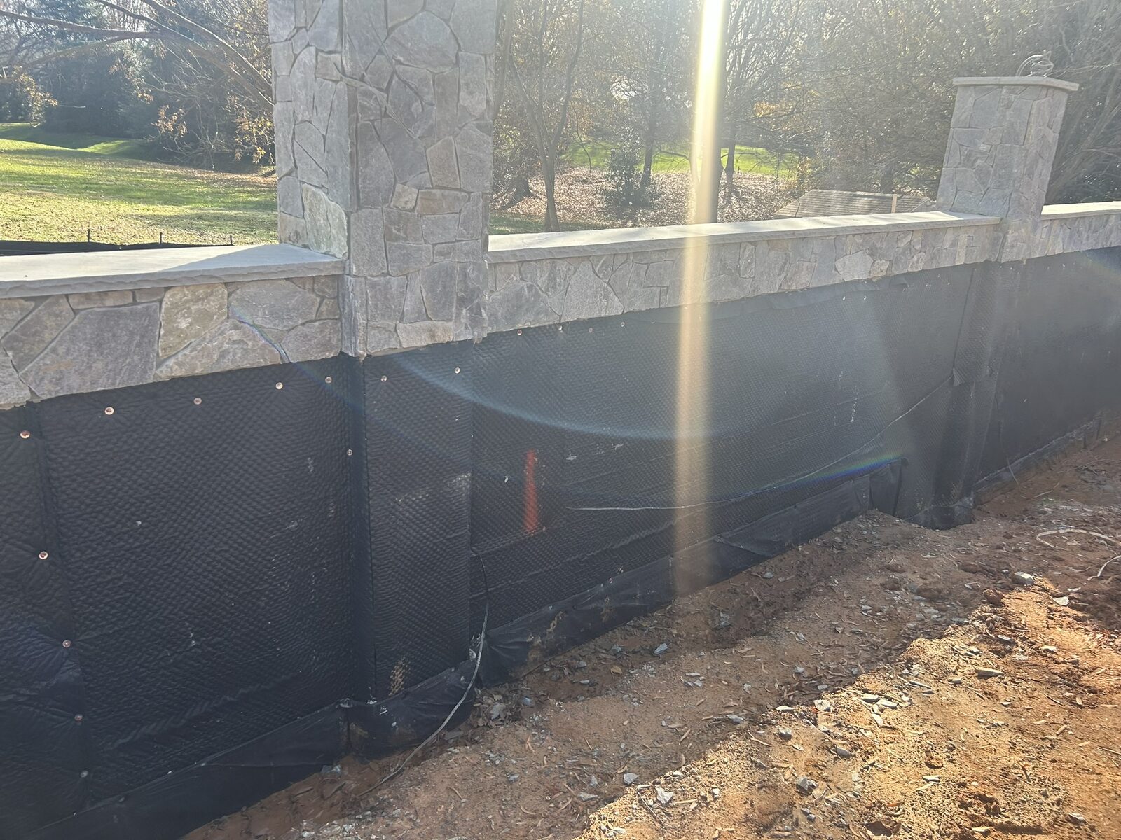 Black dimpled drainage plane and a footing drain installed along the base of the motor court retaining wall, with stone veneer and cap stone finished above.